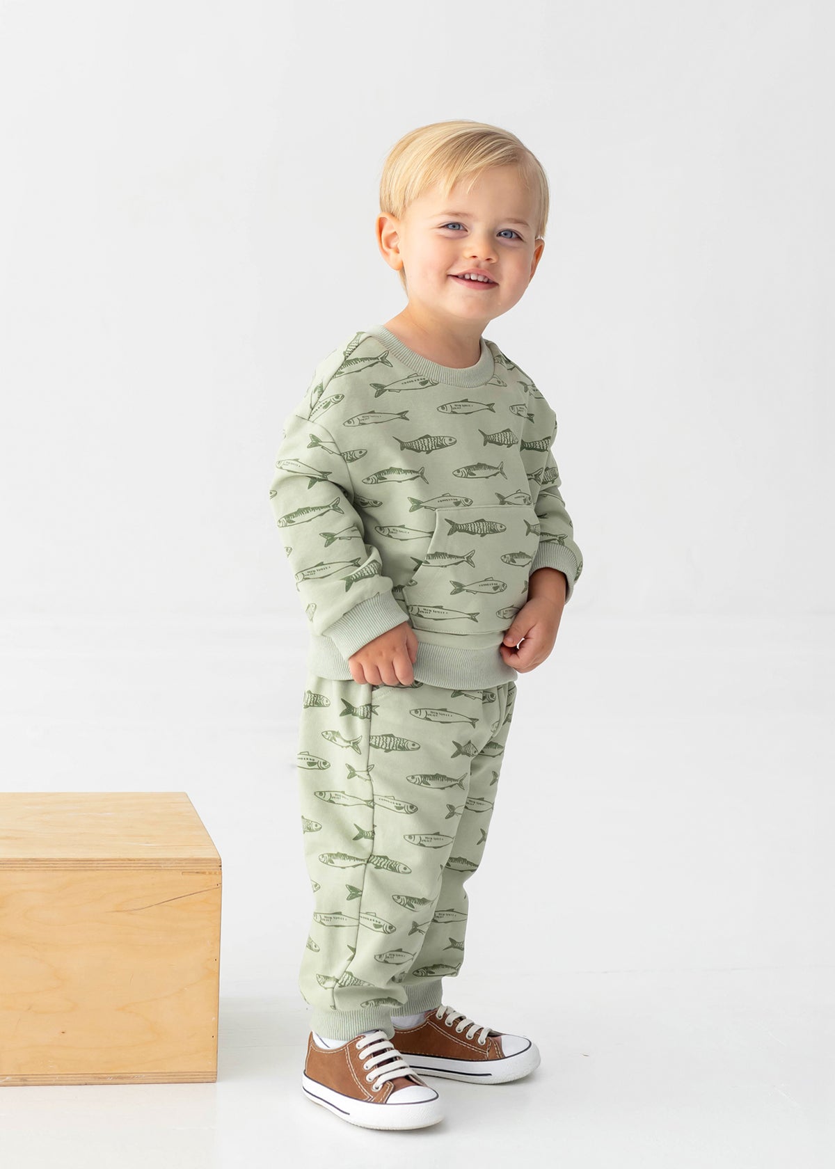 A smiling blond-haired toddler wears the Beckett and Bear Matthew Long Sleeve Fish Print Sweatshirt and Jogger Set in sage green, made from a soft cotton blend fleece, along with brown sneakers, and stands beside a wooden block against a plain white background.