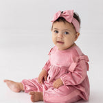 A baby sits on the floor against a plain white background, wearing the Mabel and Honey Bowtiful Blush Long Sleeve Ribbed Shoulder Bow Tie Bubble Romper with matching bows and a pink headband, looking toward the camera.
