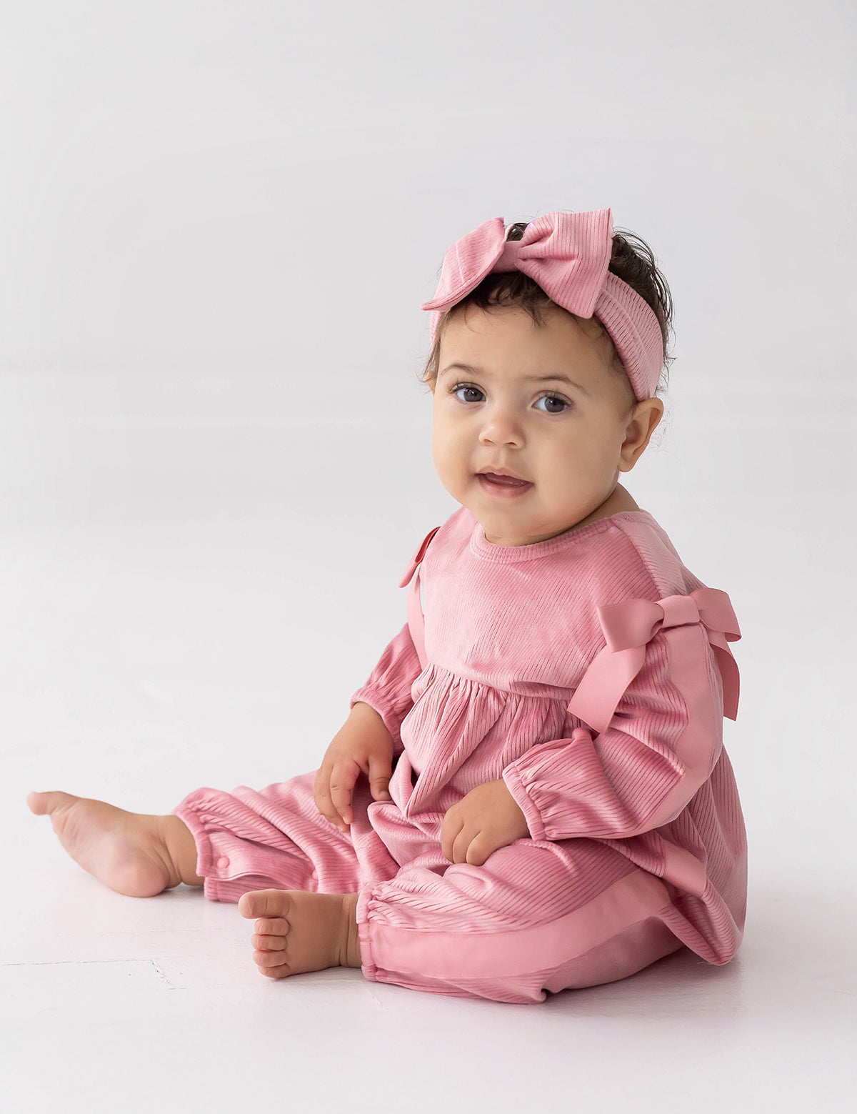 A baby sits on the floor against a plain white background, wearing the Mabel and Honey Bowtiful Blush Long Sleeve Ribbed Shoulder Bow Tie Bubble Romper with matching bows and a pink headband, looking toward the camera.