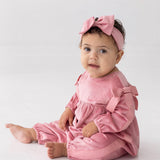 A baby sits on the floor against a plain white background, wearing the Mabel and Honey Bowtiful Blush Long Sleeve Ribbed Shoulder Bow Tie Bubble Romper with matching bows and a pink headband, looking toward the camera.