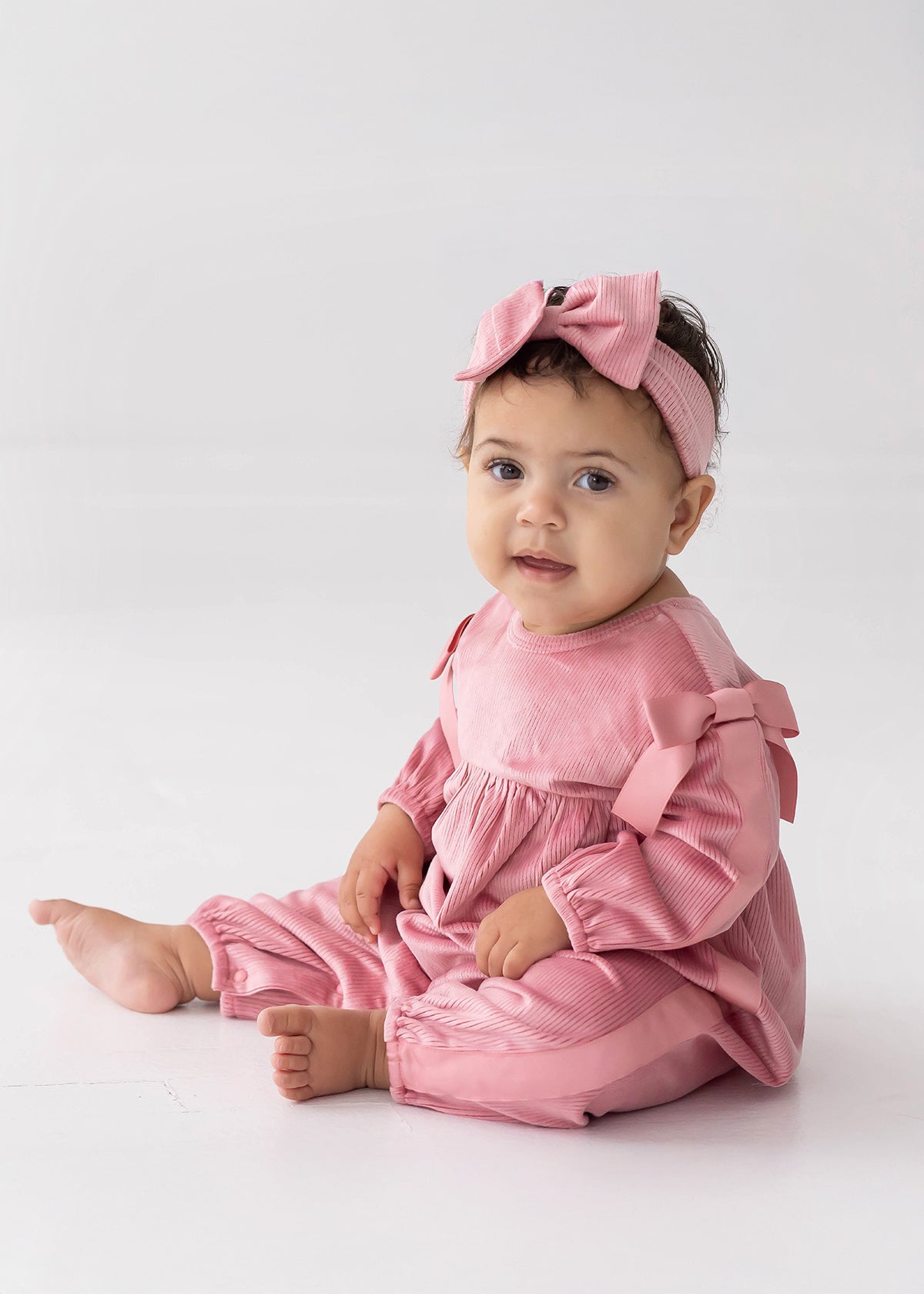A baby sits on the floor against a plain white background, wearing the Mabel and Honey Bowtiful Blush Long Sleeve Ribbed Shoulder Bow Tie Bubble Romper with matching bows and a pink headband, looking toward the camera.