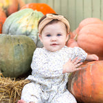 A baby wearing the Mabel and Honey Wildflower Dream Long Sleeve Floral Puff Sleeve Bubble Romper sits barefoot among large pumpkins and hay, smiling softly.