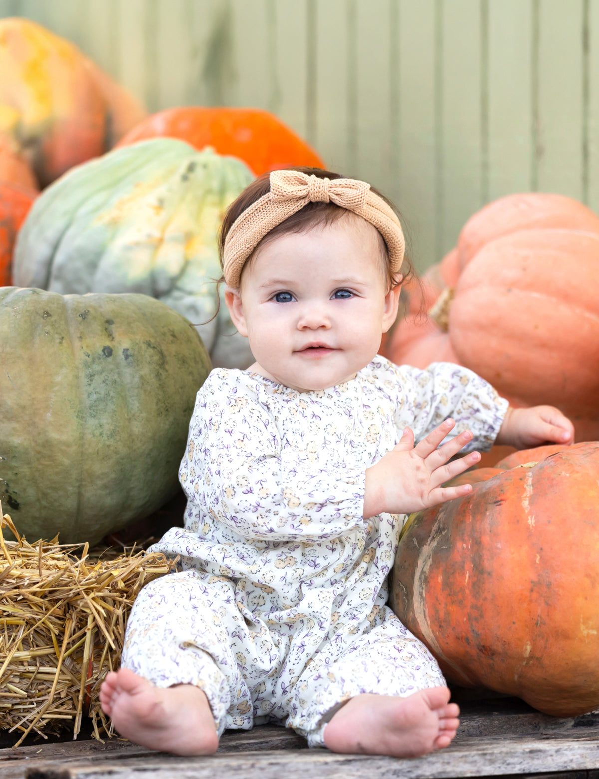 A baby wearing the Mabel and Honey Wildflower Dream Long Sleeve Floral Puff Sleeve Bubble Romper sits barefoot among large pumpkins and hay, smiling softly.