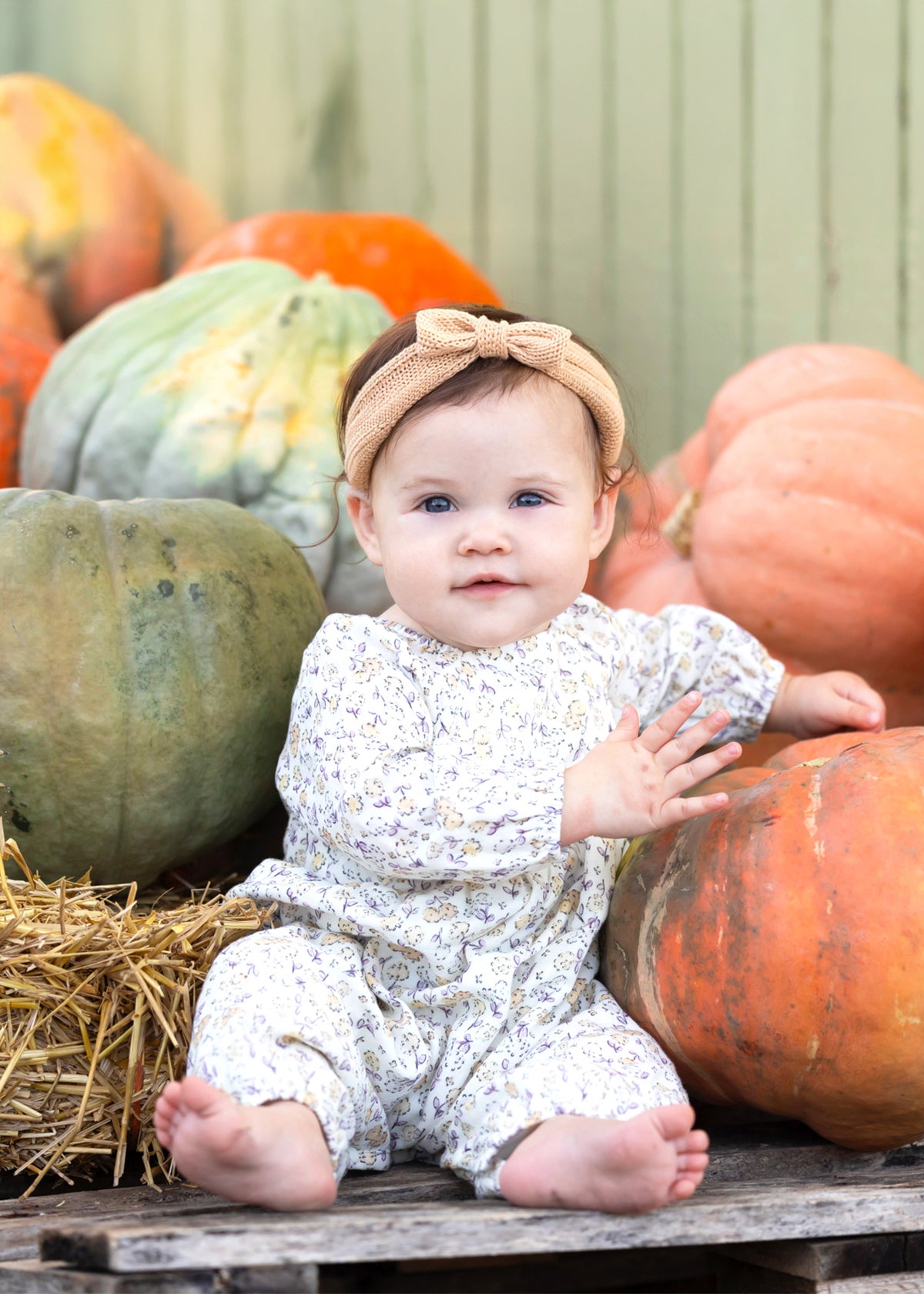 A baby wearing the Mabel and Honey Wildflower Dream Long Sleeve Floral Puff Sleeve Bubble Romper sits barefoot among large pumpkins and hay, smiling softly.