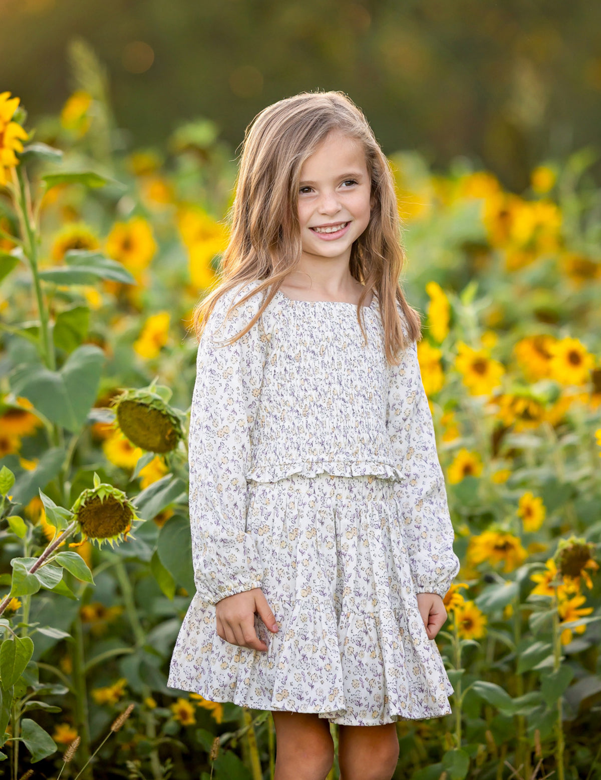A young girl with long hair stands smiling in a field of sunflowers under soft sunlight, wearing the Mabel and Honey Wildflower Dream Long Sleeve Floral Smocked Top and Skirt Set.