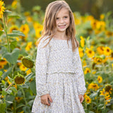 A young girl with long hair stands smiling in a field of sunflowers under soft sunlight, wearing the Mabel and Honey Wildflower Dream Long Sleeve Floral Smocked Top and Skirt Set.