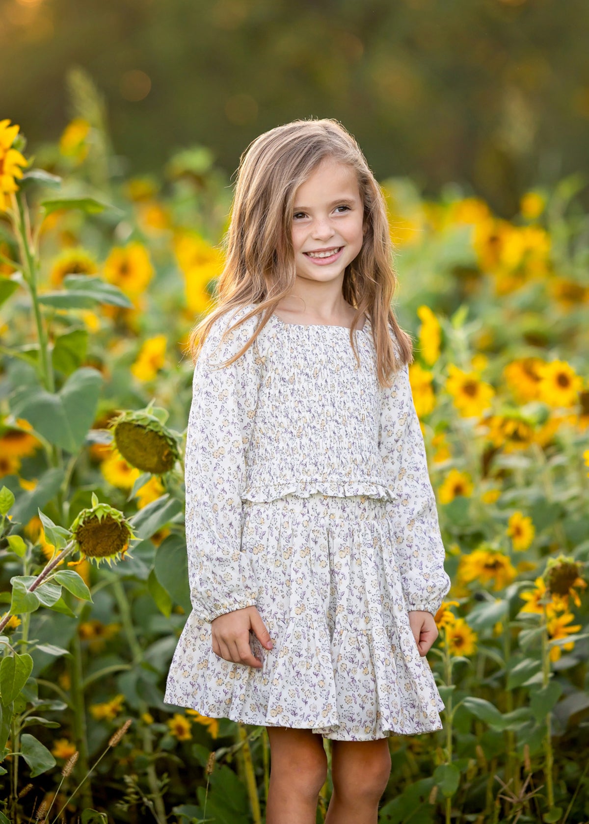 A young girl with long hair stands smiling in a field of sunflowers under soft sunlight, wearing the Mabel and Honey Wildflower Dream Long Sleeve Floral Smocked Top and Skirt Set.