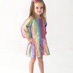 A young girl wearing the Isobella and Chloe Rainbow Party Long Sleeve Color Block Sequin Ruffle Hem Set with a matching bow stands against a white background, her hand on her hip and a slight smile on her face.