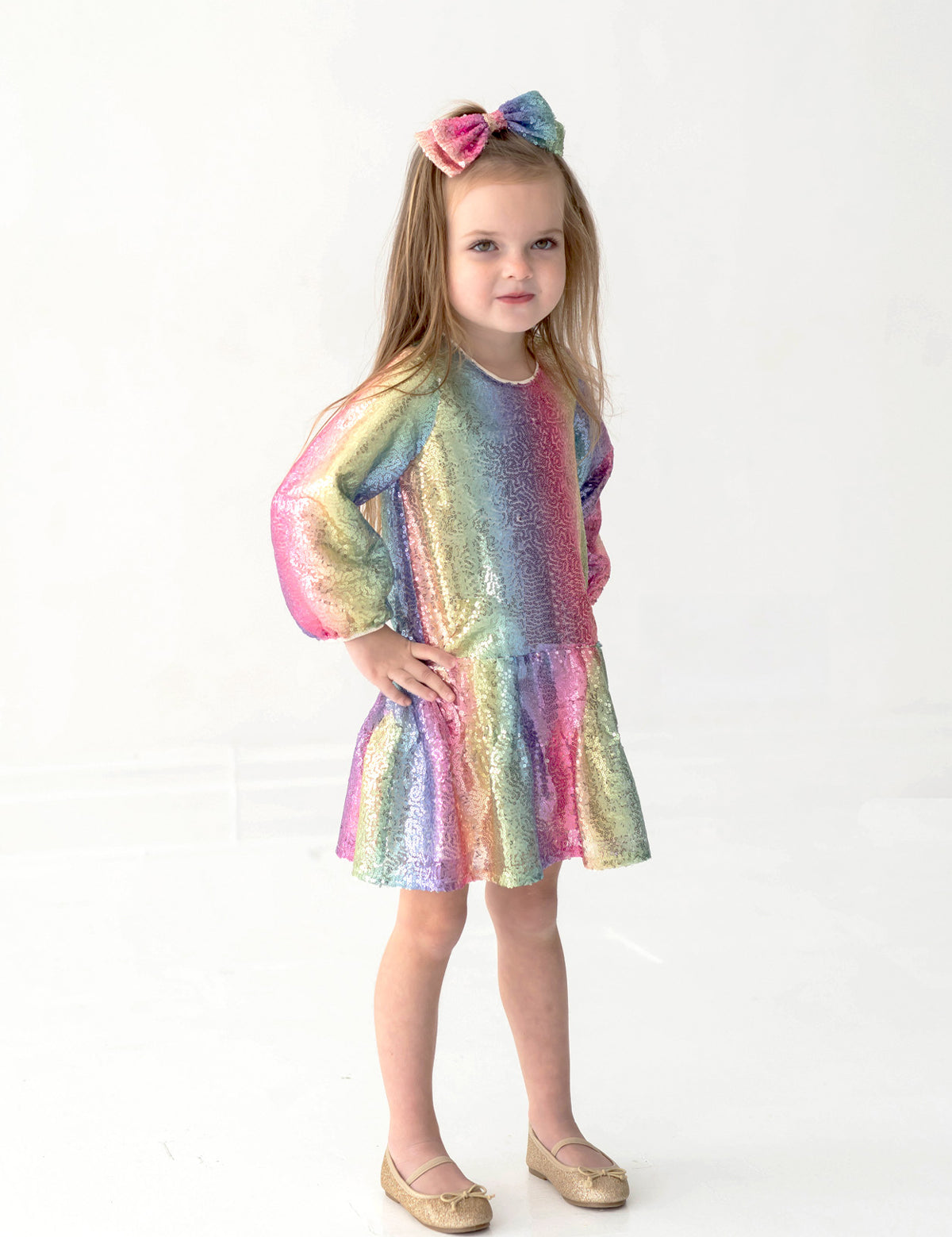 A young girl wearing the Isobella and Chloe Rainbow Party Long Sleeve Color Block Sequin Ruffle Hem Set with a matching bow stands against a white background, her hand on her hip and a slight smile on her face.