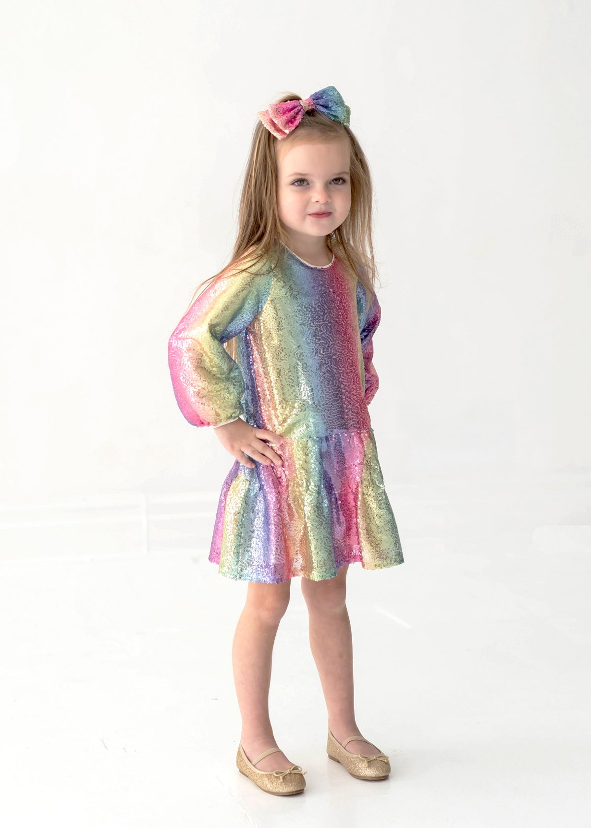 A young girl wearing the Isobella and Chloe Rainbow Party Long Sleeve Color Block Sequin Ruffle Hem Set with a matching bow stands against a white background, her hand on her hip and a slight smile on her face.
