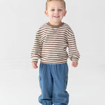 A smiling young child stands on a white background, wearing a brown and white striped long-sleeve shirt, Mabel and Honey Michael Denim Cargo Pocket Jogger Pants featuring an elastic waistband and cargo pockets, and white sneakers with dark stripes.
