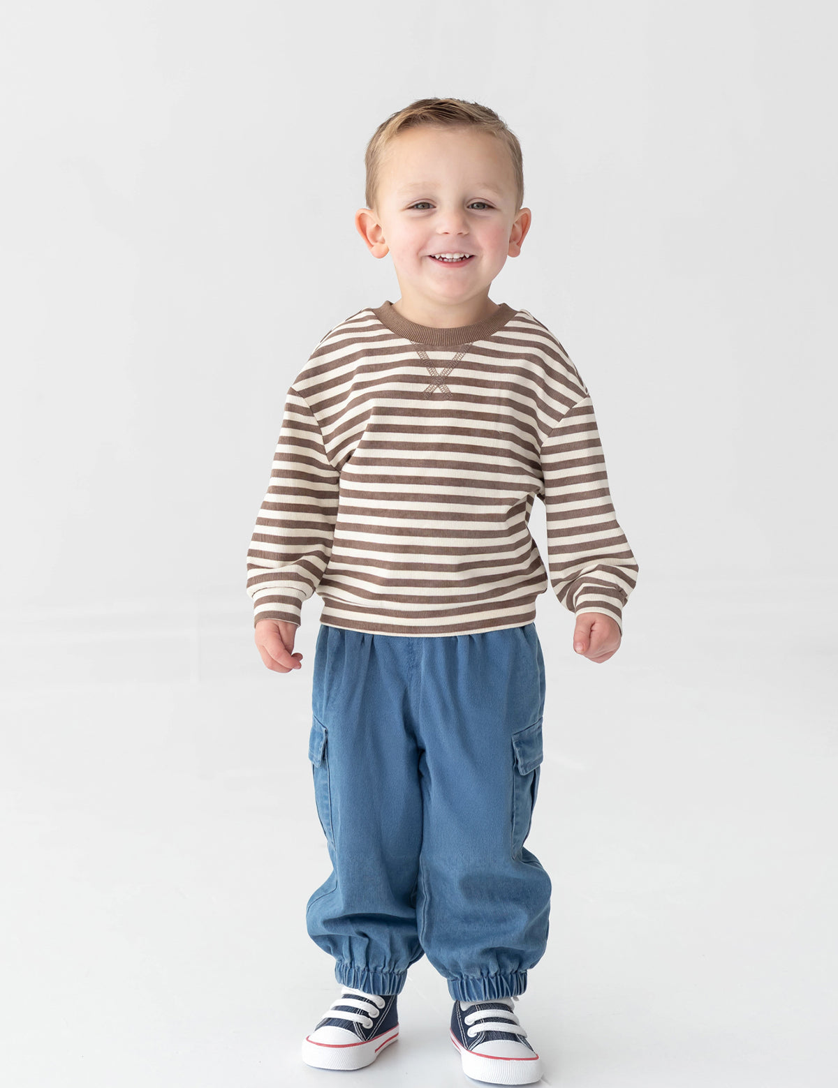 A smiling young child stands on a white background, wearing a brown and white striped long-sleeve shirt, Mabel and Honey Michael Denim Cargo Pocket Jogger Pants featuring an elastic waistband and cargo pockets, and white sneakers with dark stripes.