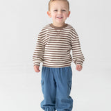 A smiling young child stands on a white background, wearing a brown and white striped long-sleeve shirt, Mabel and Honey Michael Denim Cargo Pocket Jogger Pants featuring an elastic waistband and cargo pockets, and white sneakers with dark stripes.