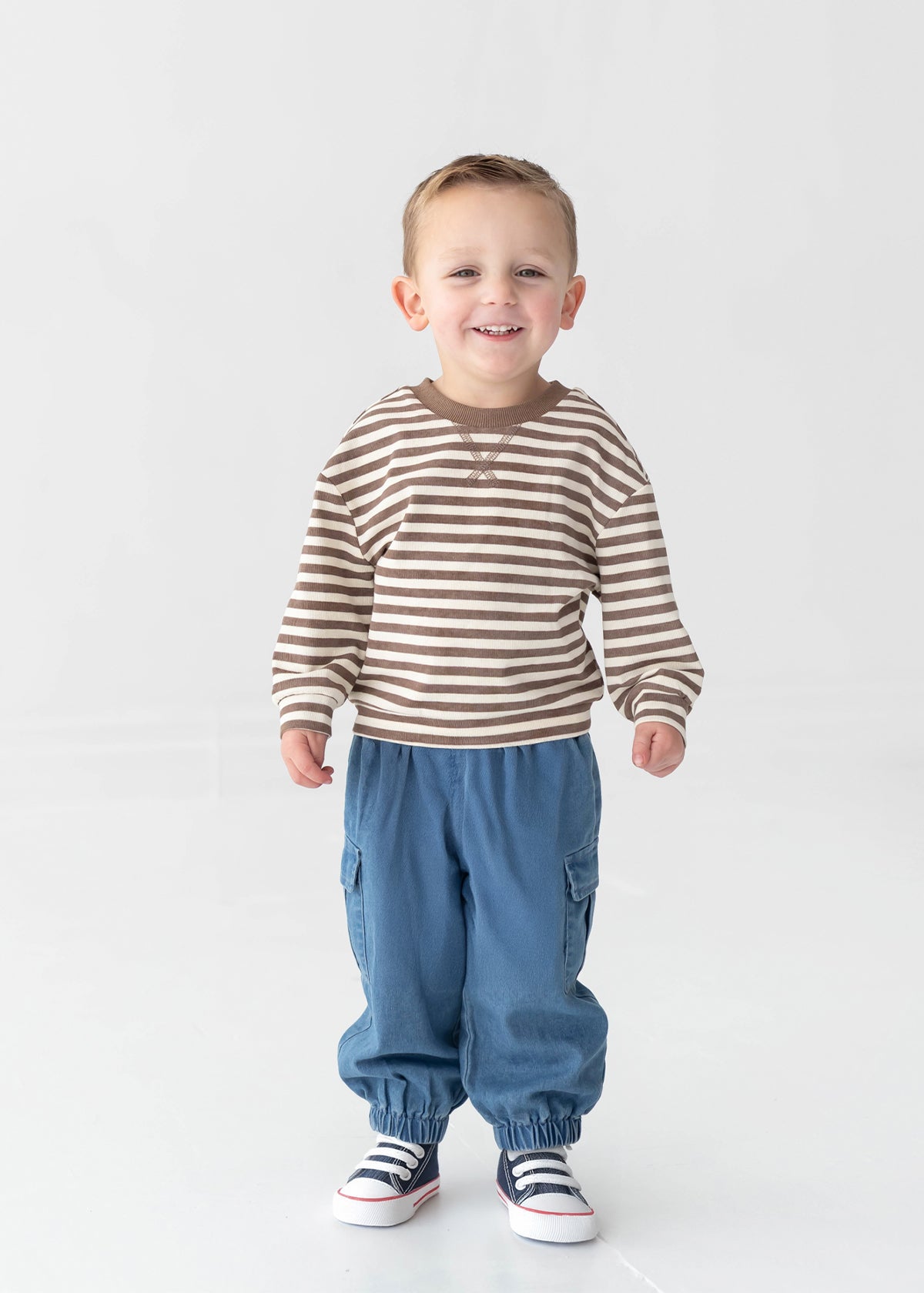 A smiling young child stands on a white background, wearing a brown and white striped long-sleeve shirt, Mabel and Honey Michael Denim Cargo Pocket Jogger Pants featuring an elastic waistband and cargo pockets, and white sneakers with dark stripes.