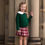 A young girl with long wavy hair stands smiling beside large stone columns, dressed in a Mabel and Honey Evergreen Long Sleeve Ribbed Knit Sweater layered over a white-collared shirt, paired with a red plaid skirt and white boots.