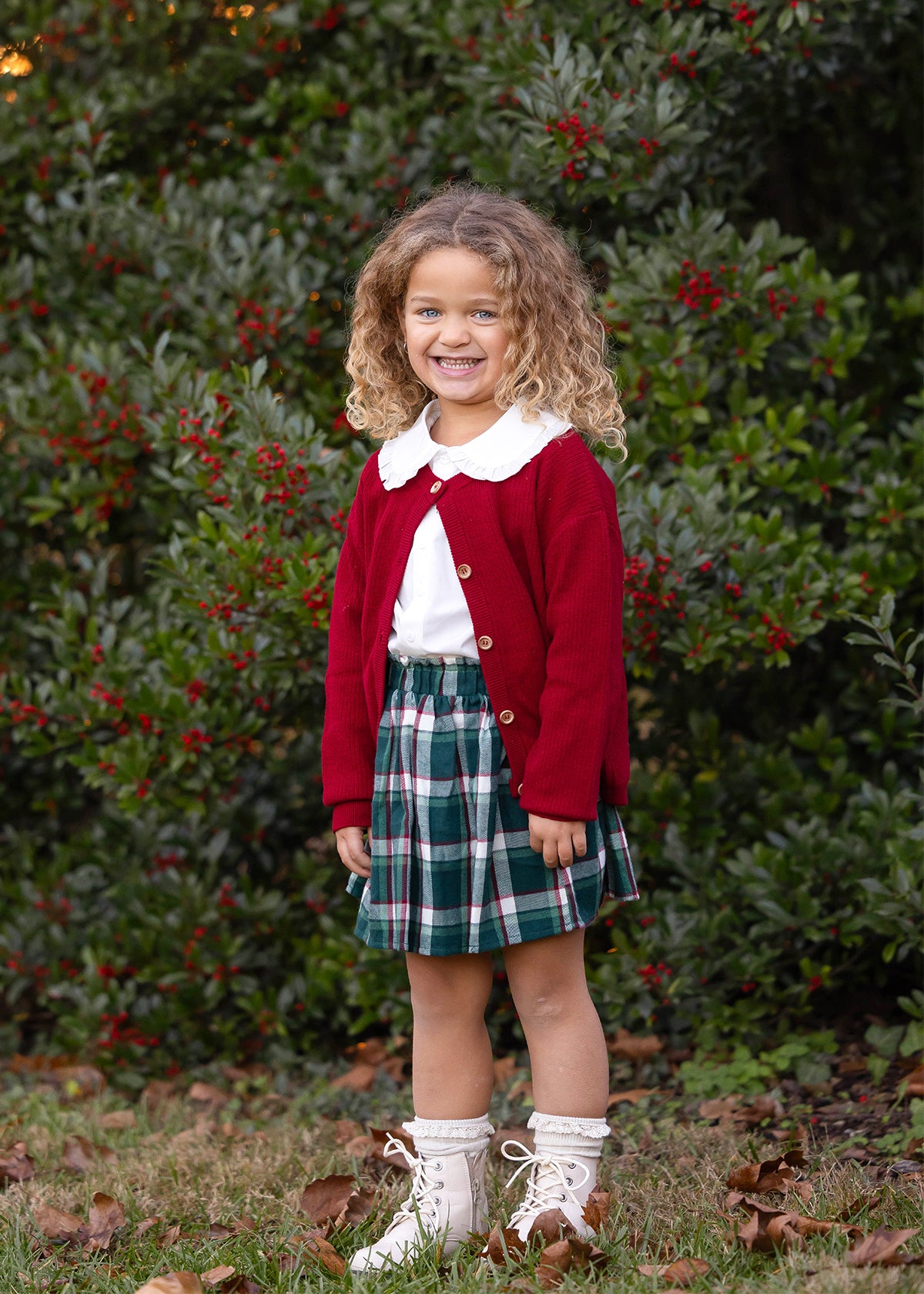 A young girl with curly hair smiles outdoors in front of a leafy bush with red berries, wearing a red cardigan, white shirt, and the Mabel and Honey Evergreen Pleated Elastic Waist Flannel Skirt with Straight Hem, paired with boots.