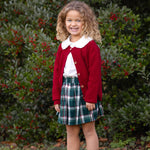 A young girl with curly hair stands smiling outdoors, wearing the Mabel and Honey Winterberry Ribbed Long Sleeve Button Front Cardigan Sweater in red over a white shirt, paired with a plaid skirt, white socks, and boots. Green bushes dotted with red berries fill the background.