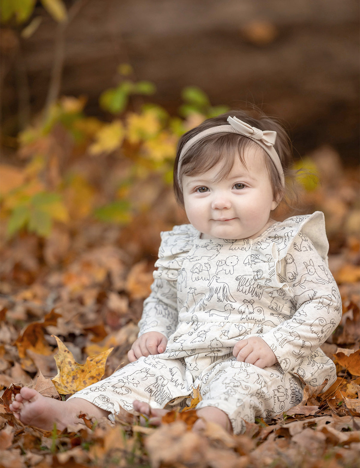A baby in the Mabel and Honey Doggie Doodles Long Sleeve Print Ruffle Collar Straight Leg Romper, complete with a matching headband and crafted from a soft cotton blend, sits barefoot among autumn leaves, smiling softly.