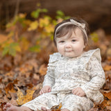 A baby in the Mabel and Honey Doggie Doodles Long Sleeve Print Ruffle Collar Straight Leg Romper, complete with a matching headband and crafted from a soft cotton blend, sits barefoot among autumn leaves, smiling softly.