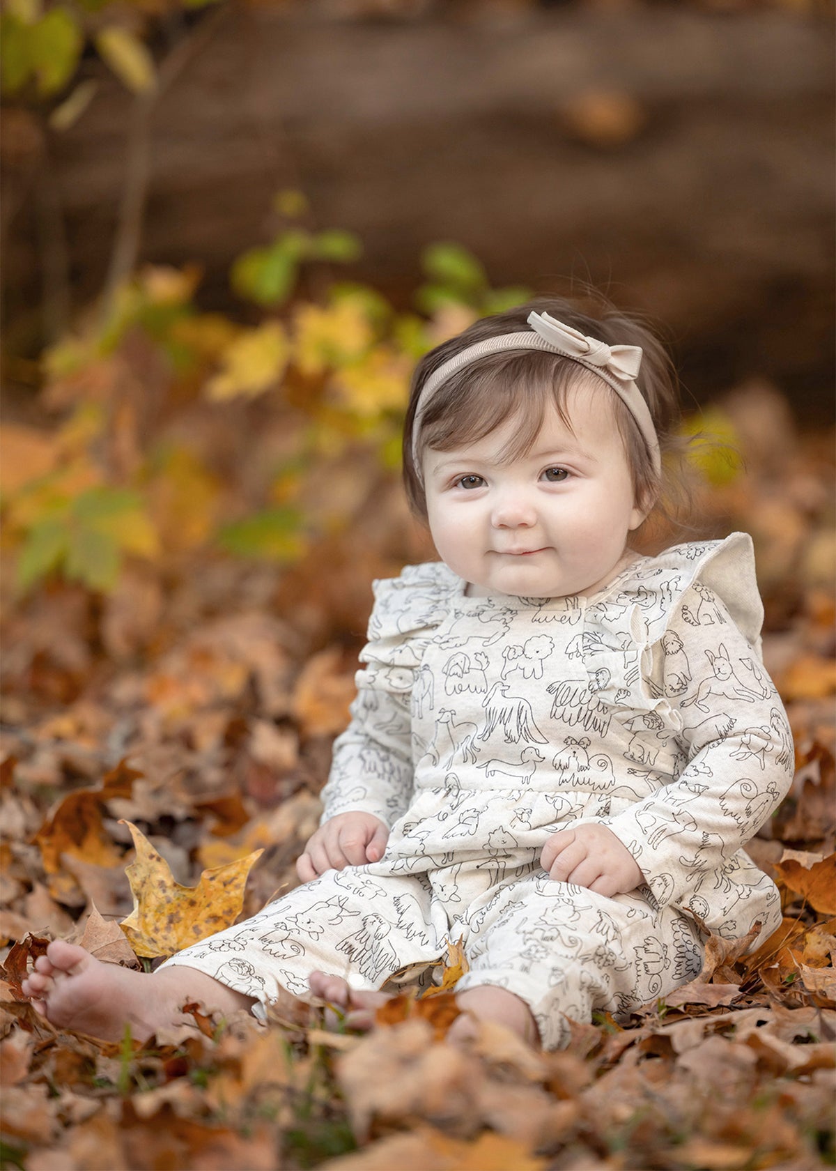 A baby in the Mabel and Honey Doggie Doodles Long Sleeve Print Ruffle Collar Straight Leg Romper, complete with a matching headband and crafted from a soft cotton blend, sits barefoot among autumn leaves, smiling softly.