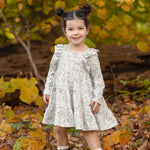 A young girl with dark hair in two buns smiles while standing on fallen leaves, wearing the Mabel and Honey Doggie Doodles Long Sleeve Print Ruffle Trim Tiered Dress and black boots. Autumn trees with yellow leaves are in the background.