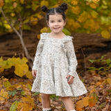 A young girl with dark hair in two buns smiles while standing on fallen leaves, wearing the Mabel and Honey Doggie Doodles Long Sleeve Print Ruffle Trim Tiered Dress and black boots. Autumn trees with yellow leaves are in the background.