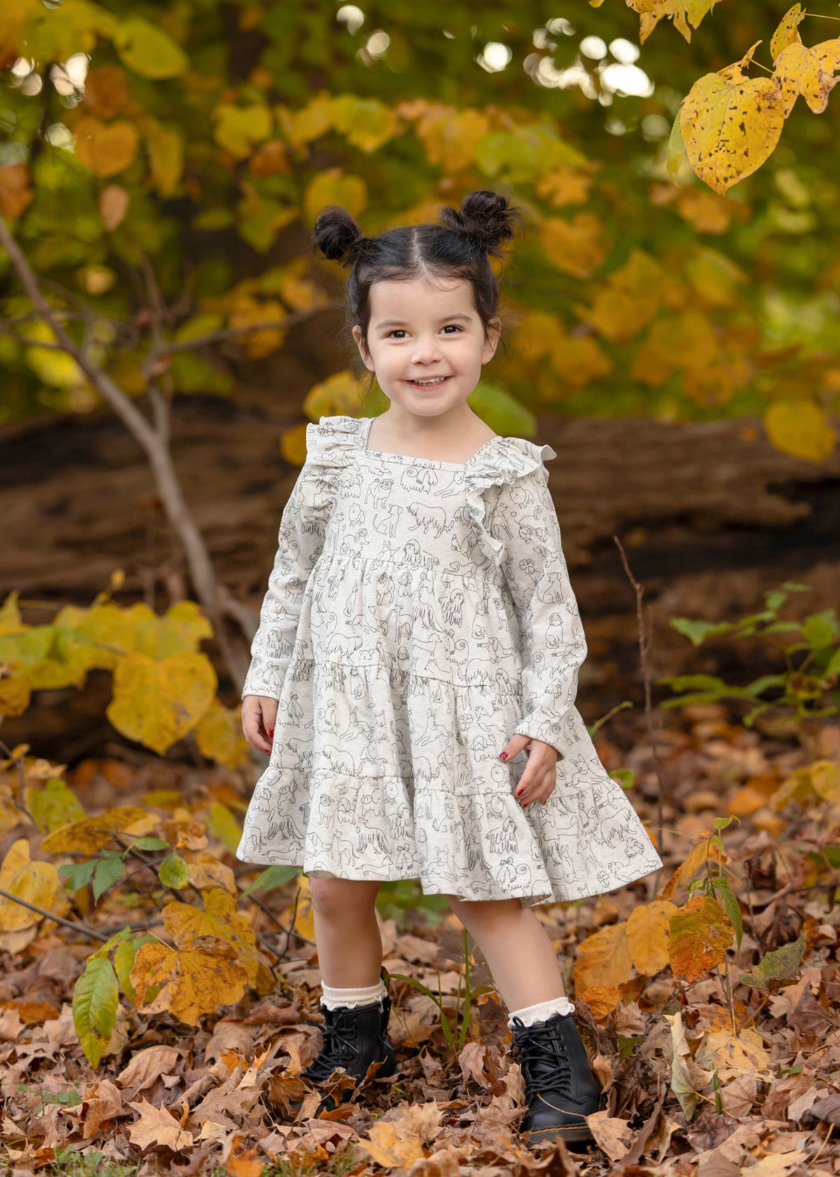 A young girl with dark hair in two buns smiles while standing on fallen leaves, wearing the Mabel and Honey Doggie Doodles Long Sleeve Print Ruffle Trim Tiered Dress and black boots. Autumn trees with yellow leaves are in the background.