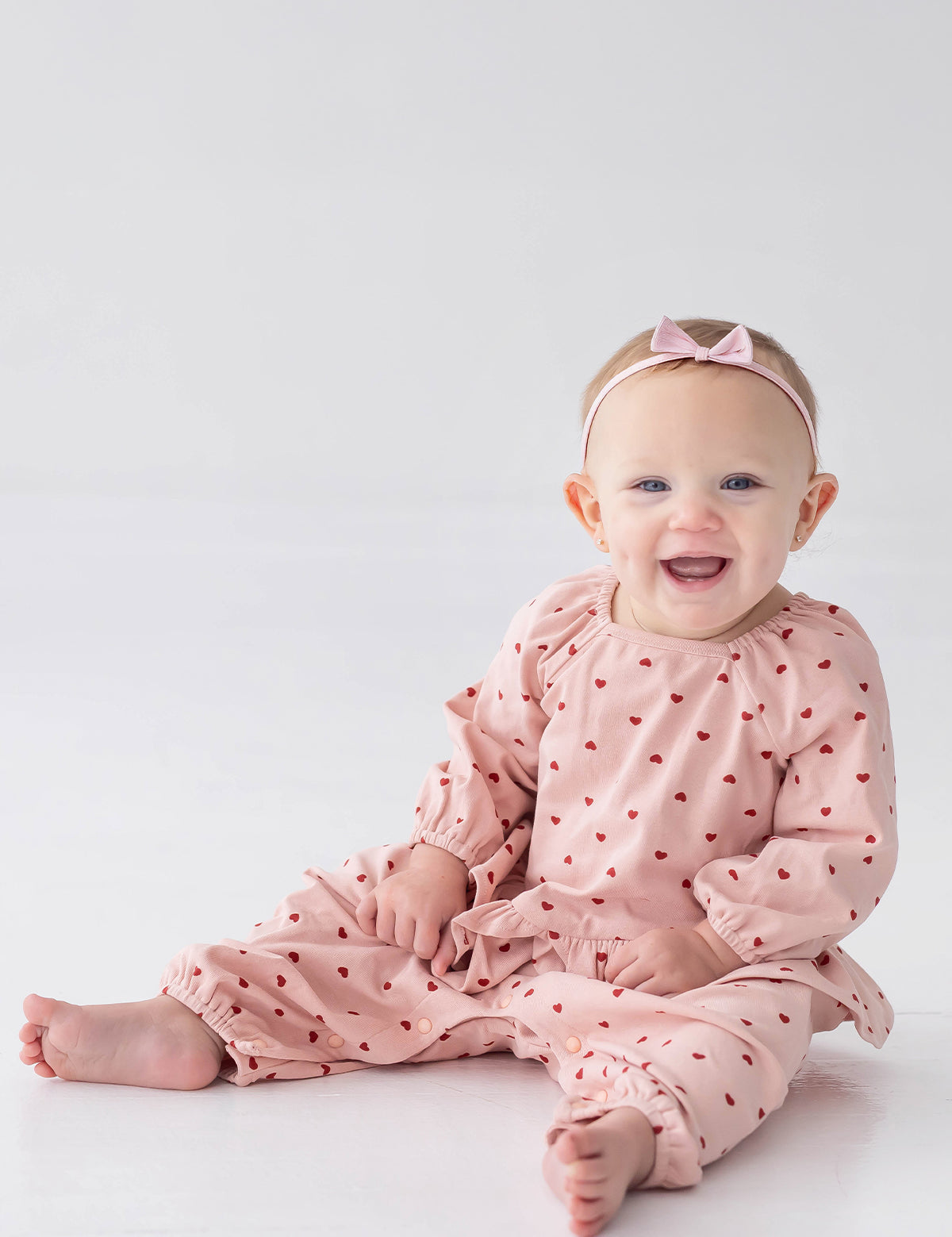 A smiling baby sits barefoot on a white floor against a plain white background, wearing the Mabel and Honey Love me Lots Long Sleeve Heart Print Ruffle Waist Romper in blush pink with heart prints and a coordinating pink bow headband.