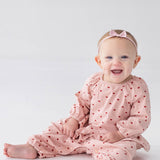 A smiling baby sits barefoot on a white floor against a plain white background, wearing the Mabel and Honey Love me Lots Long Sleeve Heart Print Ruffle Waist Romper in blush pink with heart prints and a coordinating pink bow headband.