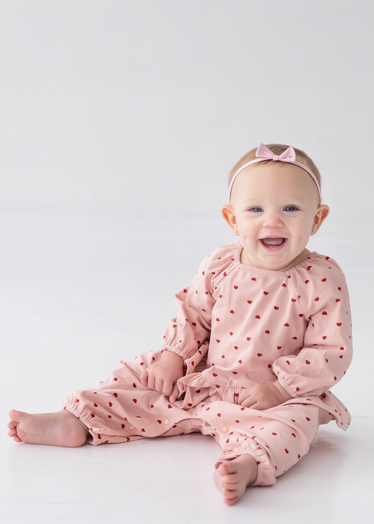 A smiling baby sits barefoot on a white floor against a plain white background, wearing the Mabel and Honey Love me Lots Long Sleeve Heart Print Ruffle Waist Romper in blush pink with heart prints and a coordinating pink bow headband.