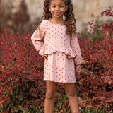 A young girl with curly hair stands outdoors, smiling. She wears the Love me Lots Long Sleeve Heart Print Ruffle Waist Dress by Mabel and Honey, paired with brown boots, surrounded by red-leaved bushes and fallen leaves.