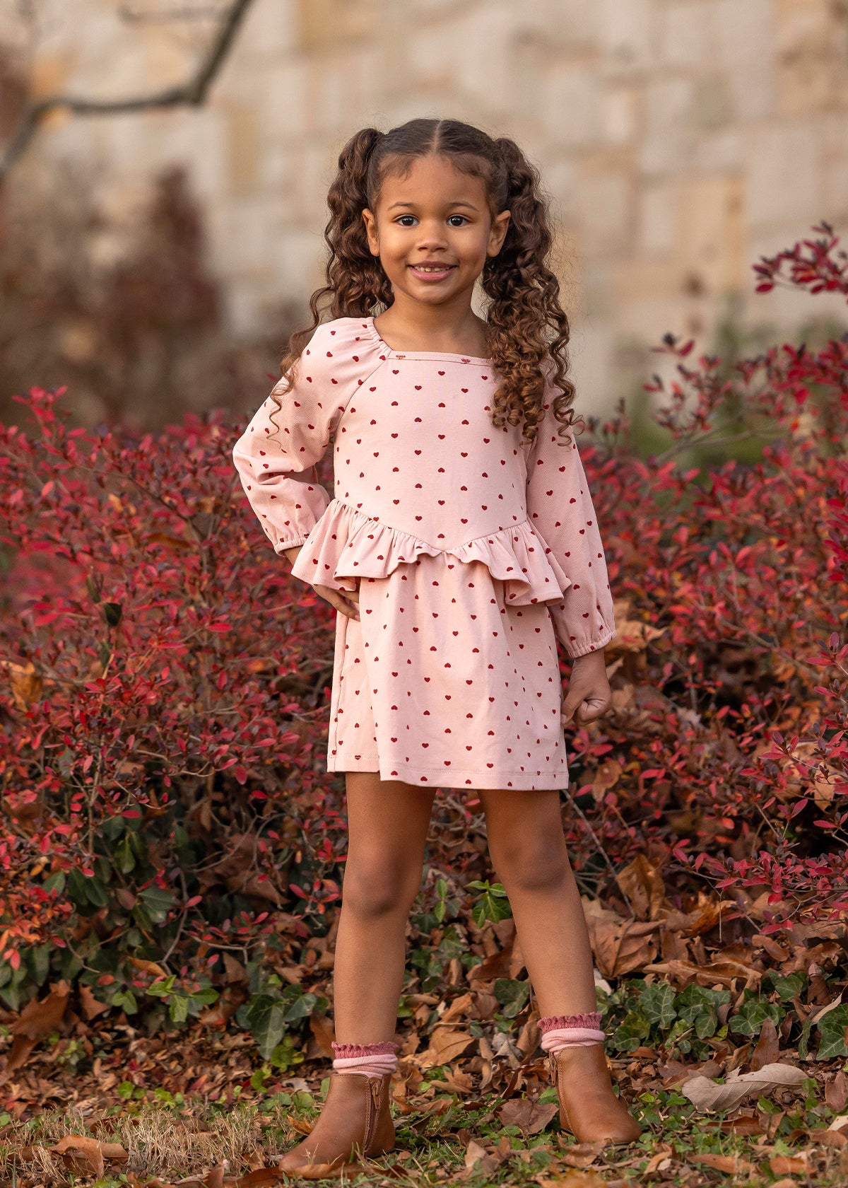 A young girl with curly hair stands outdoors, smiling. She wears the Love me Lots Long Sleeve Heart Print Ruffle Waist Dress by Mabel and Honey, paired with brown boots, surrounded by red-leaved bushes and fallen leaves.