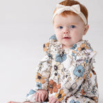 A baby with red hair, wearing a white headband with a bow and the Mabel and Honey Autumn Blooms Long Sleeve Floral Ruffle Trim Straight Leg Romper, sits on a white floor against a light background, looking toward the camera.