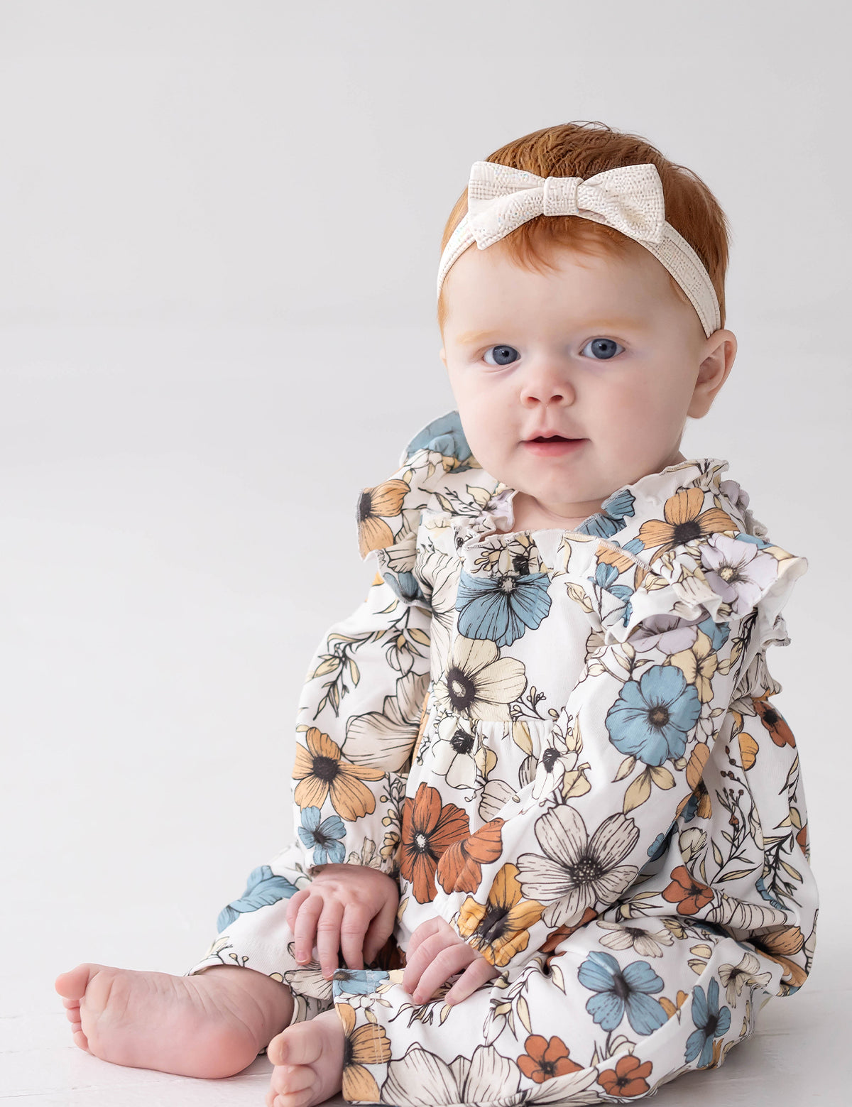 A baby with red hair, wearing a white headband with a bow and the Mabel and Honey Autumn Blooms Long Sleeve Floral Ruffle Trim Straight Leg Romper, sits on a white floor against a light background, looking toward the camera.