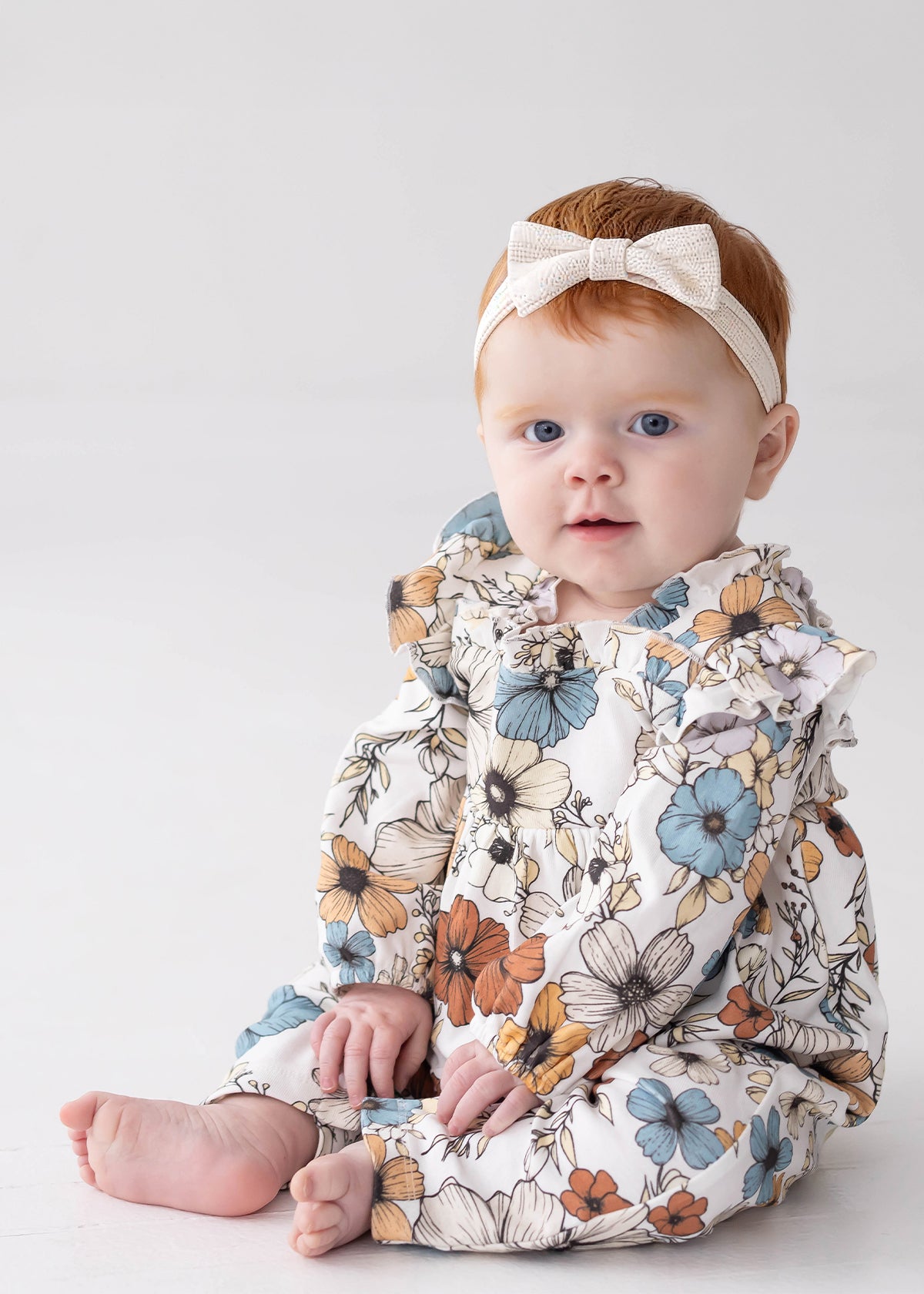 A baby with red hair, wearing a white headband with a bow and the Mabel and Honey Autumn Blooms Long Sleeve Floral Ruffle Trim Straight Leg Romper, sits on a white floor against a light background, looking toward the camera.