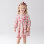A young child with light hair stands and smiles against a plain light background, wearing the Mabel and Honey “Caroline” Long Sleeve Floral Tiered Contrast Piping Dress, paired with white boots and a pink headband.