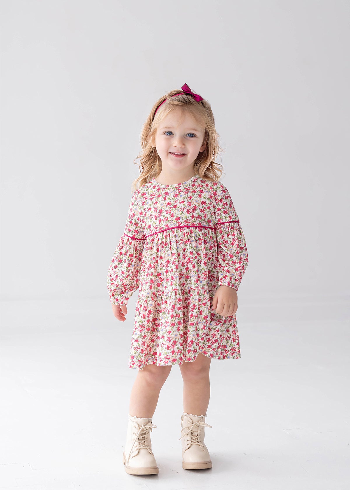 A young child with light hair stands and smiles against a plain light background, wearing the Mabel and Honey “Caroline” Long Sleeve Floral Tiered Contrast Piping Dress, paired with white boots and a pink headband.