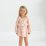 A young girl with blonde hair smiles while standing in a light room. She wears the Mabel and Honey Honey Petals Long Sleeve Floral Ruffle Top and Legging Set, featuring a soft knit floral top with ruffles, peach leggings, and white sneakers—the perfect fall outfit for girls.