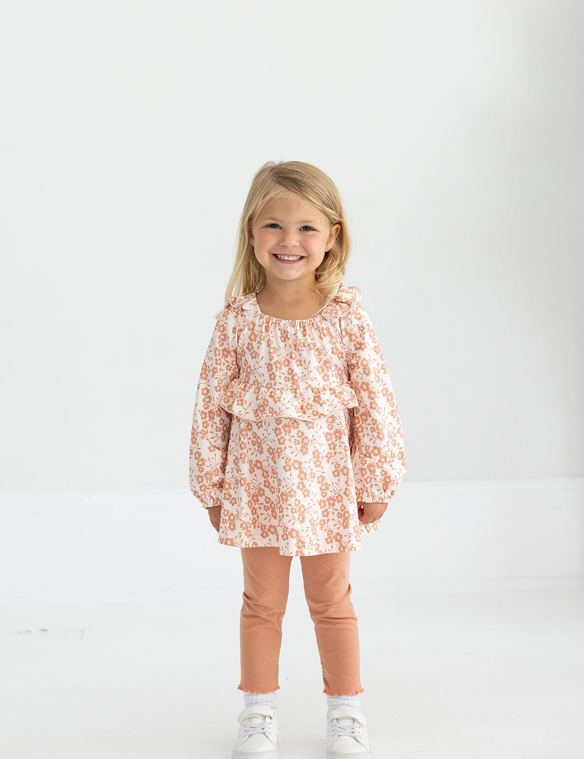 A young girl with blonde hair smiles while standing in a light room. She wears the Mabel and Honey Honey Petals Long Sleeve Floral Ruffle Top and Legging Set, featuring a soft knit floral top with ruffles, peach leggings, and white sneakers—the perfect fall outfit for girls.