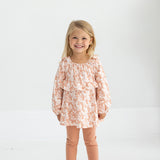 A young girl with blonde hair smiles while standing in a light room. She wears the Mabel and Honey Honey Petals Long Sleeve Floral Ruffle Top and Legging Set, featuring a soft knit floral top with ruffles, peach leggings, and white sneakers—the perfect fall outfit for girls.