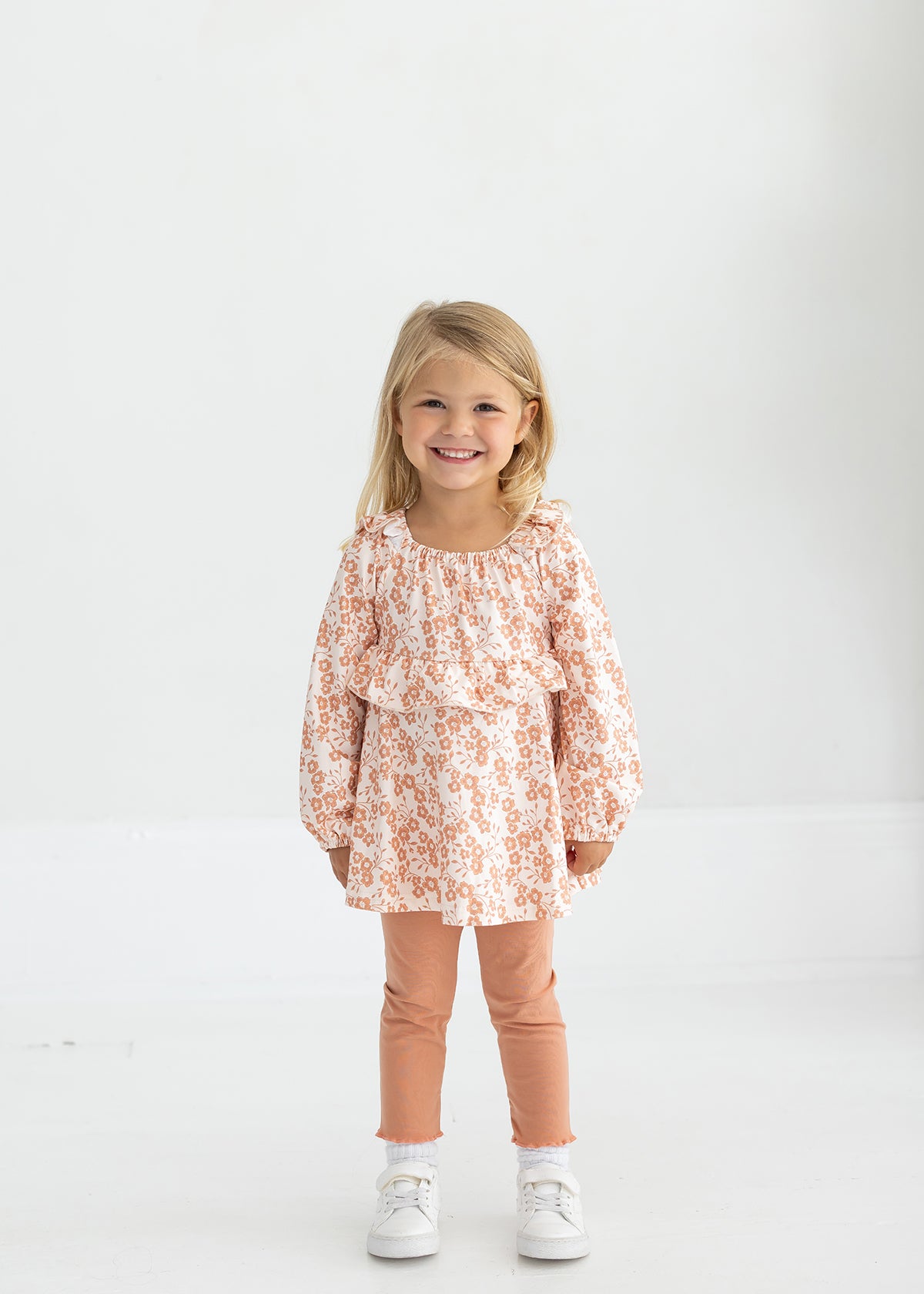 A young girl with blonde hair smiles while standing in a light room. She wears the Mabel and Honey Honey Petals Long Sleeve Floral Ruffle Top and Legging Set, featuring a soft knit floral top with ruffles, peach leggings, and white sneakers—the perfect fall outfit for girls.