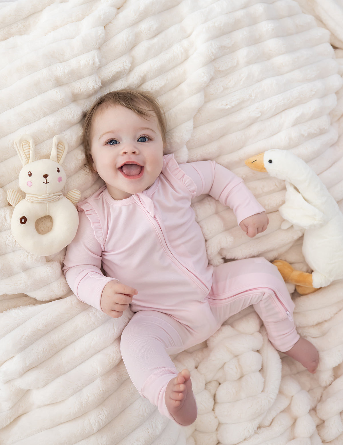 A smiling baby in the Mabel and Honey Amelia Rose Long Sleeve Solid Ruffle Shoulder Convertible Romper in pink lies on a soft, cream-colored blanket, surrounded by a plush bunny toy and a plush white goose.