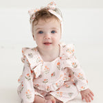 A baby with blue eyes and light brown hair, dressed in the Mabel and Honey Amelia Rose Long Sleeve Floral Ruffle Shoulder Bubble Romper with a matching headband, sits on a white floor and smiles at the camera.