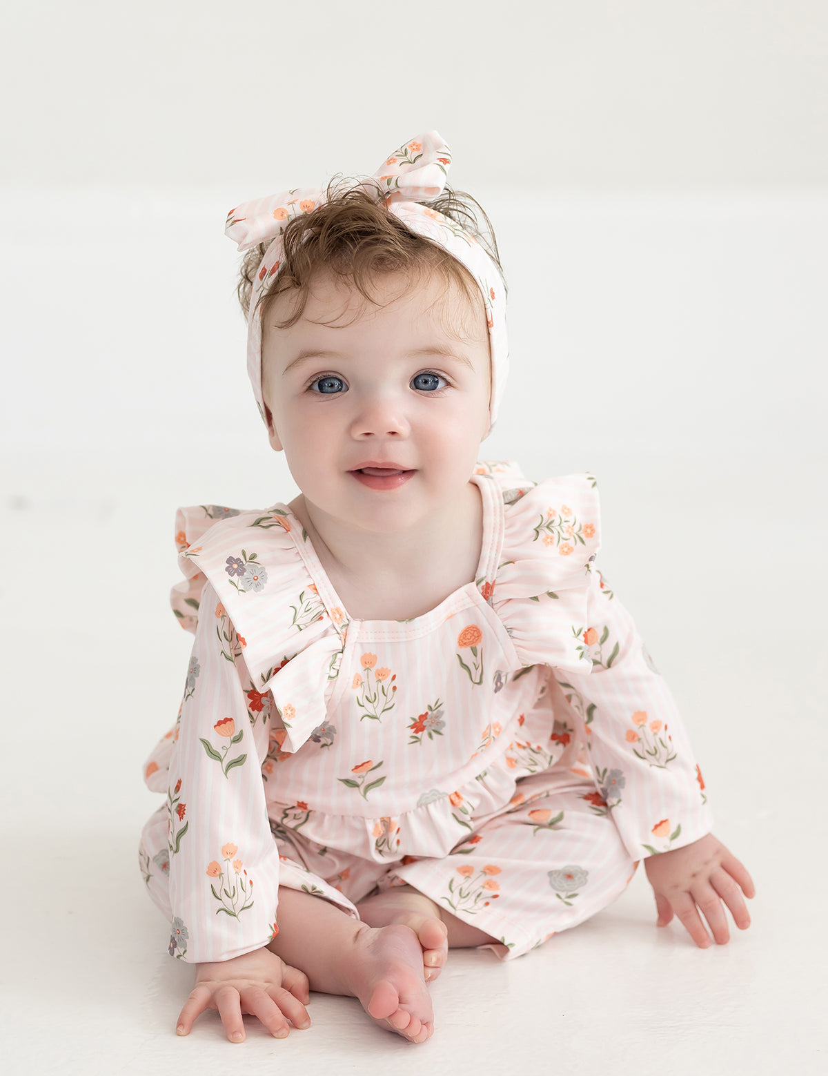 A baby with blue eyes and light brown hair, dressed in the Mabel and Honey Amelia Rose Long Sleeve Floral Ruffle Shoulder Bubble Romper with a matching headband, sits on a white floor and smiles at the camera.