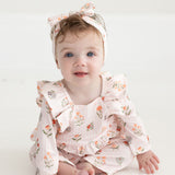 A baby with blue eyes and light brown hair, dressed in the Mabel and Honey Amelia Rose Long Sleeve Floral Ruffle Shoulder Bubble Romper with a matching headband, sits on a white floor and smiles at the camera.