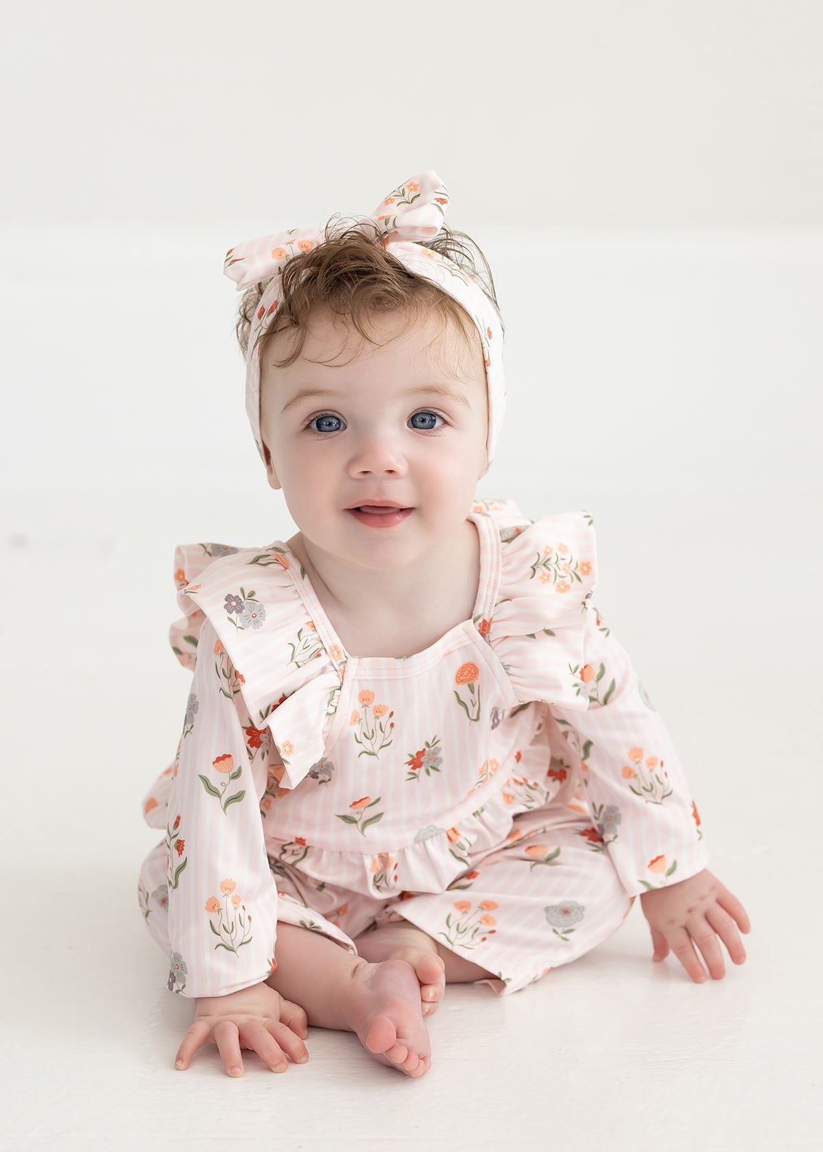 A baby with blue eyes and light brown hair, dressed in the Mabel and Honey Amelia Rose Long Sleeve Floral Ruffle Shoulder Bubble Romper with a matching headband, sits on a white floor and smiles at the camera.