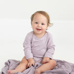 A baby with light brown hair, dressed in the Mabel and Honey Soft Meadow Long Sleeve Solid Scalloped Trim Bodysuit made from a soft cotton blend in lavender, sits smiling on a coordinating purple blanket atop a white floor.