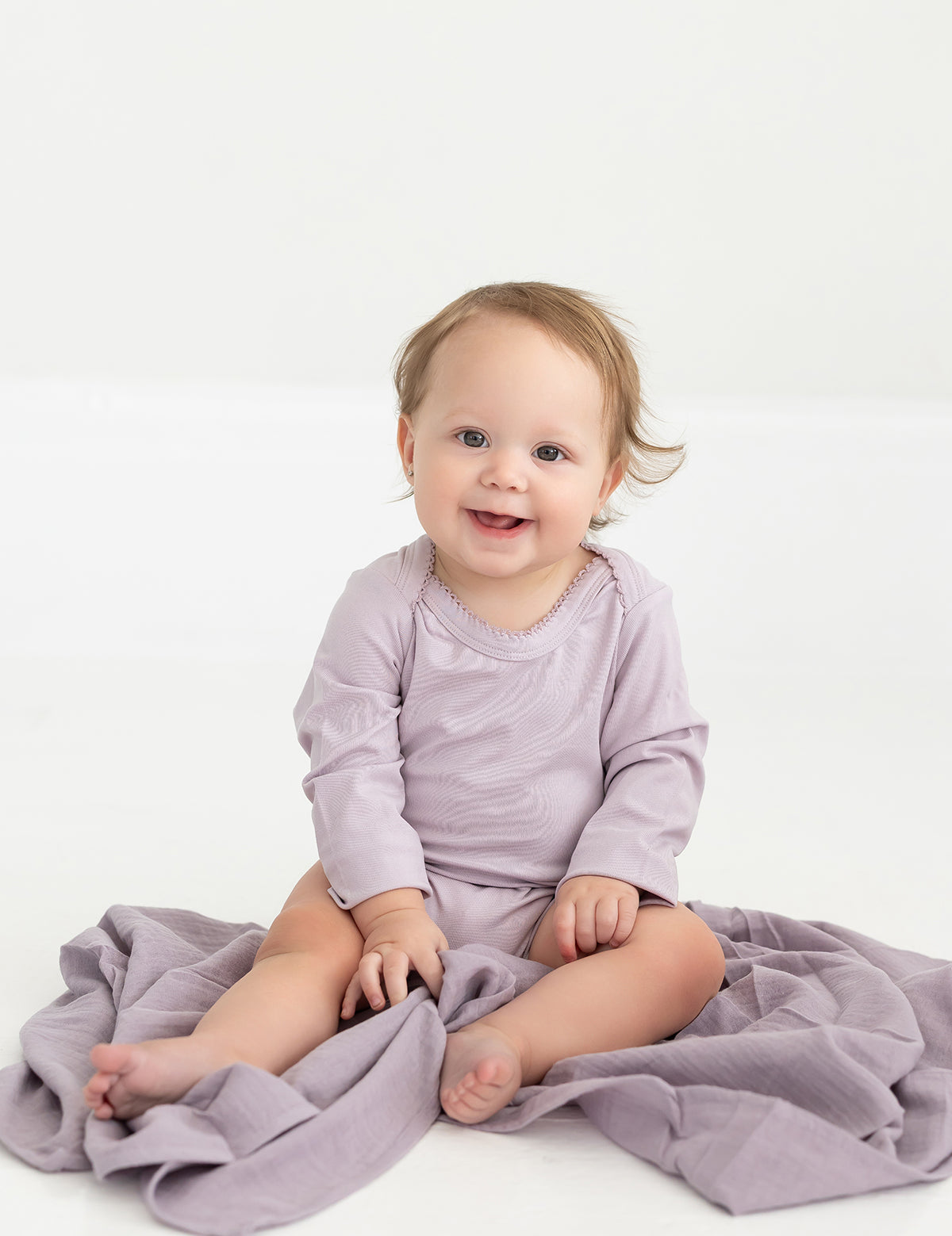 A baby with light brown hair, dressed in the Mabel and Honey Soft Meadow Long Sleeve Solid Scalloped Trim Bodysuit made from a soft cotton blend in lavender, sits smiling on a coordinating purple blanket atop a white floor.