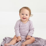 A baby with light brown hair, dressed in the Mabel and Honey Soft Meadow Long Sleeve Solid Scalloped Trim Bodysuit made from a soft cotton blend in lavender, sits smiling on a coordinating purple blanket atop a white floor.