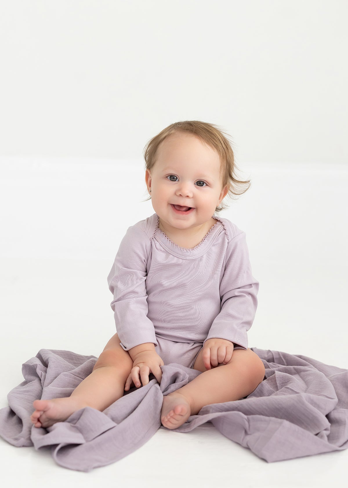 A baby with light brown hair, dressed in the Mabel and Honey Soft Meadow Long Sleeve Solid Scalloped Trim Bodysuit made from a soft cotton blend in lavender, sits smiling on a coordinating purple blanket atop a white floor.